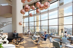 A modern hotel lobby with large windows, a staircase, and a seating area with tables and chairs.