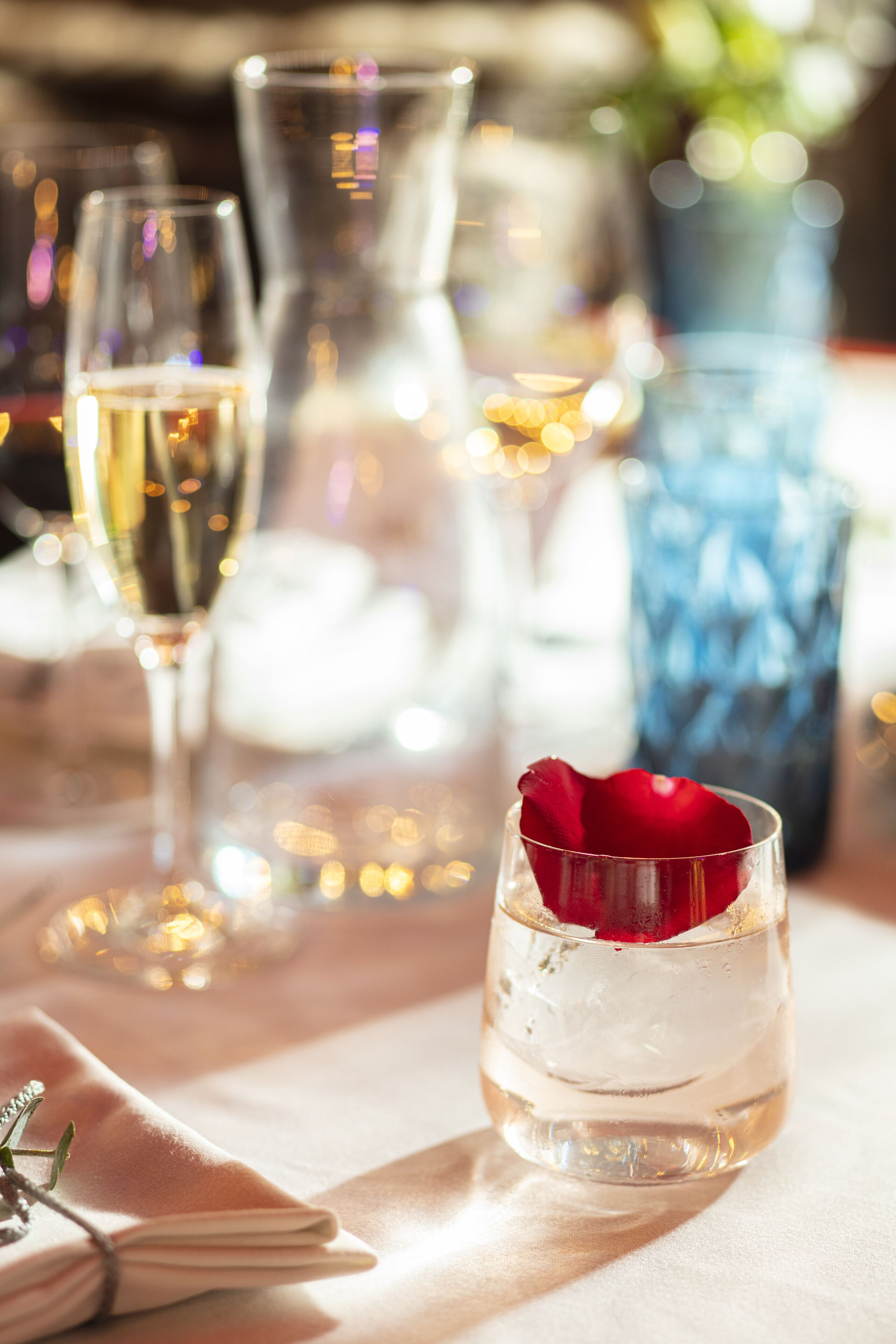 Un vaso con una rosa roja y hielo, un vaso transparente con un líquido dorado, y un florero azul al fondo.
