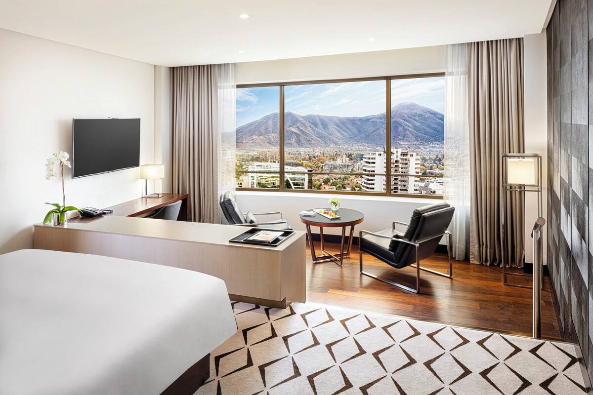 A hotel room with a large bed, a desk, a chair, a small table, and a view of mountains and a city.