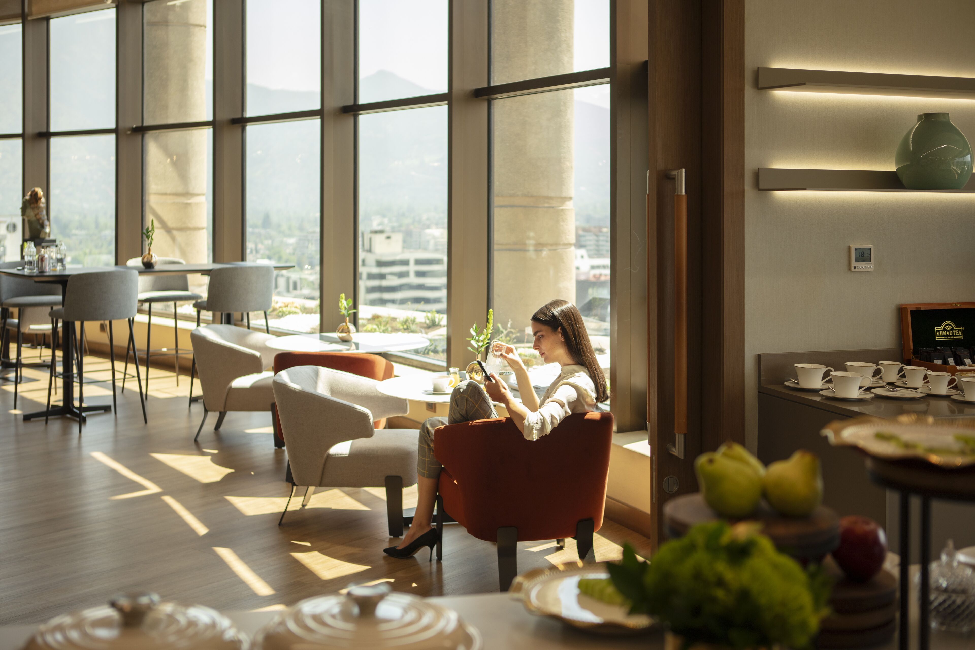 A modern hotel room with large windows, a seating area with chairs, a dining table, and a fruit display.