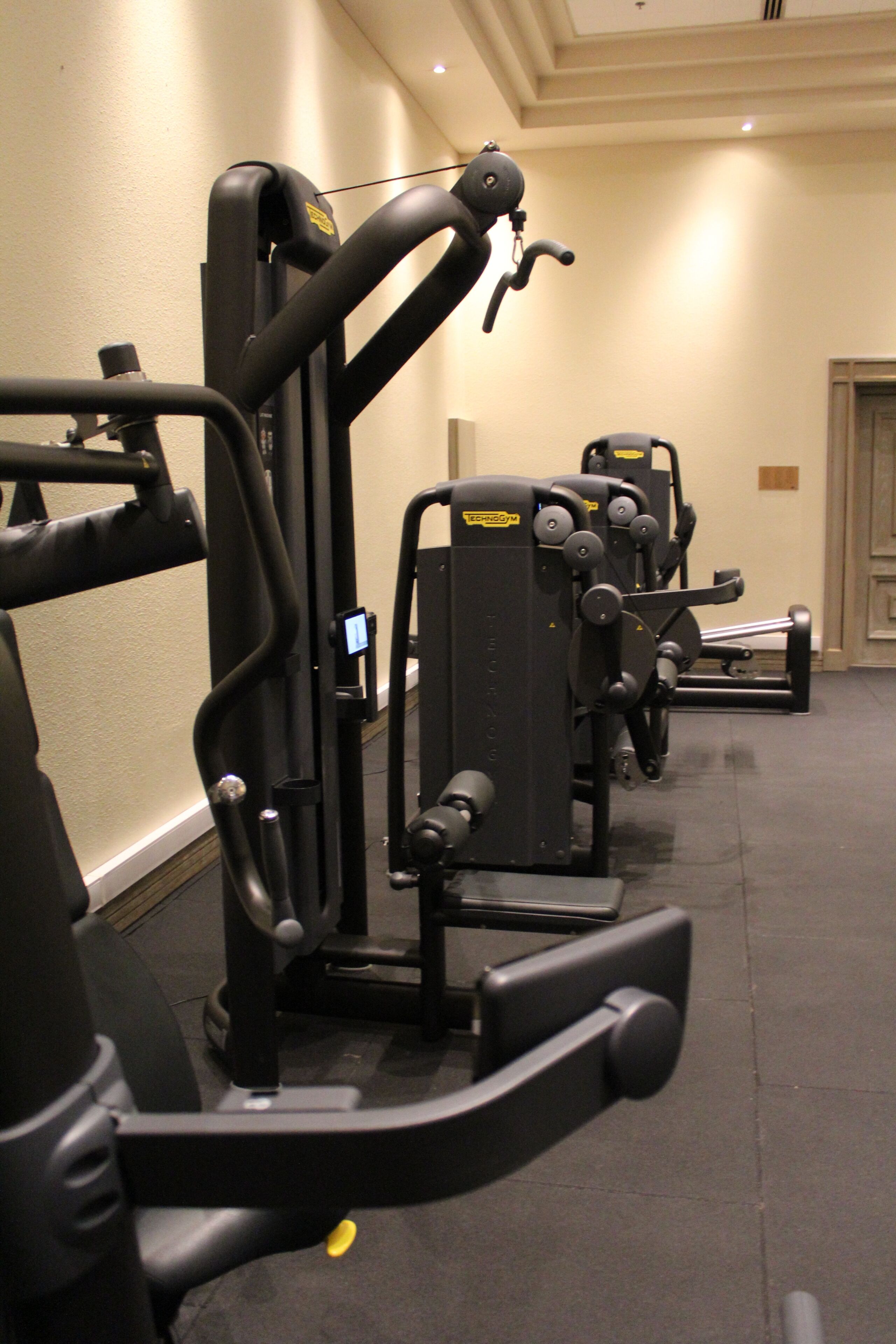A gym with various exercise machines, a wall-mounted television, and a door in the background.