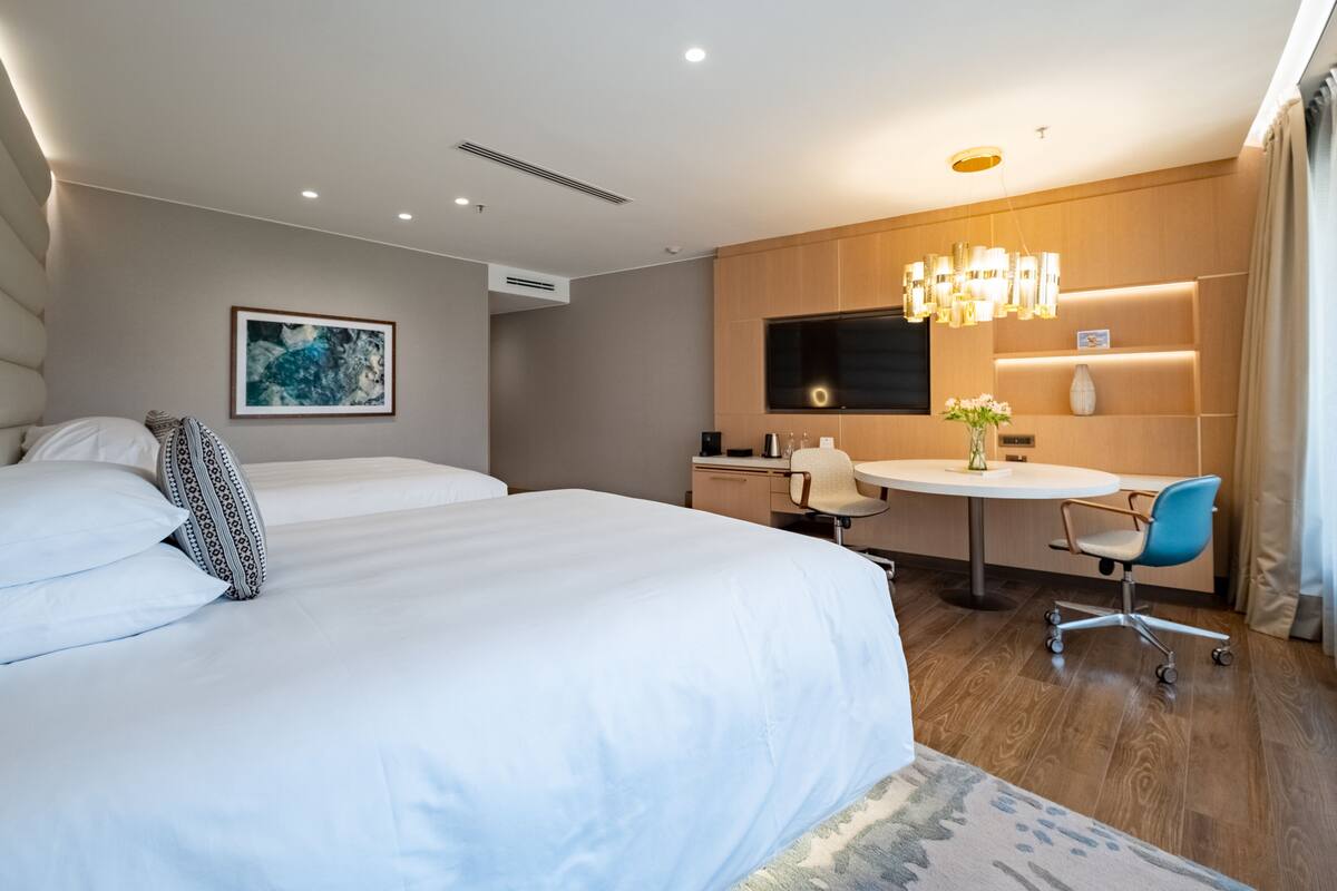 A hotel room with a large bed, a desk, a chair, a TV, and a small table with a vase.
