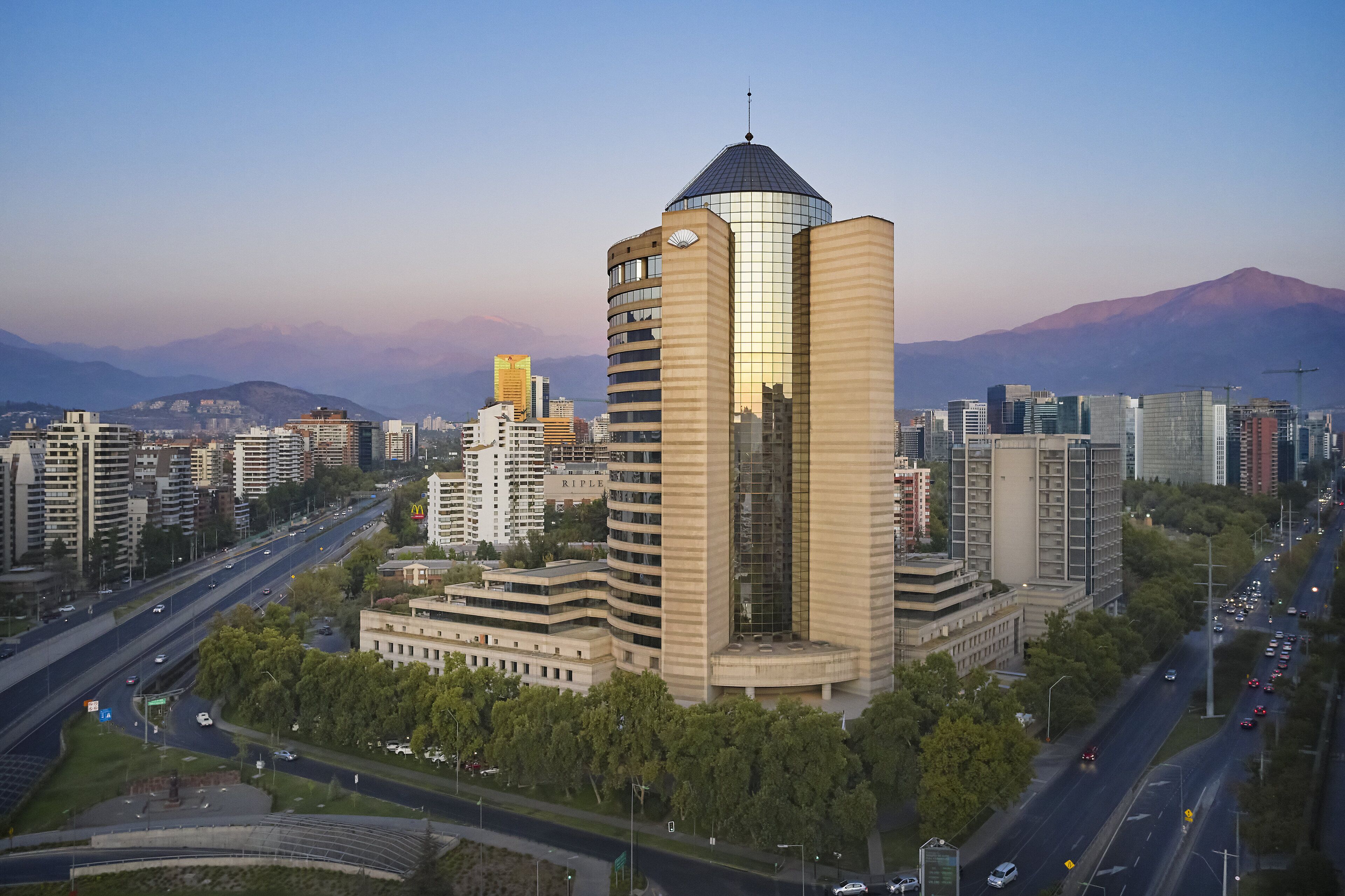 A modern building with a unique cylindrical tower and a dome, surrounded by other high-rise buildings and a mountainous backdrop.