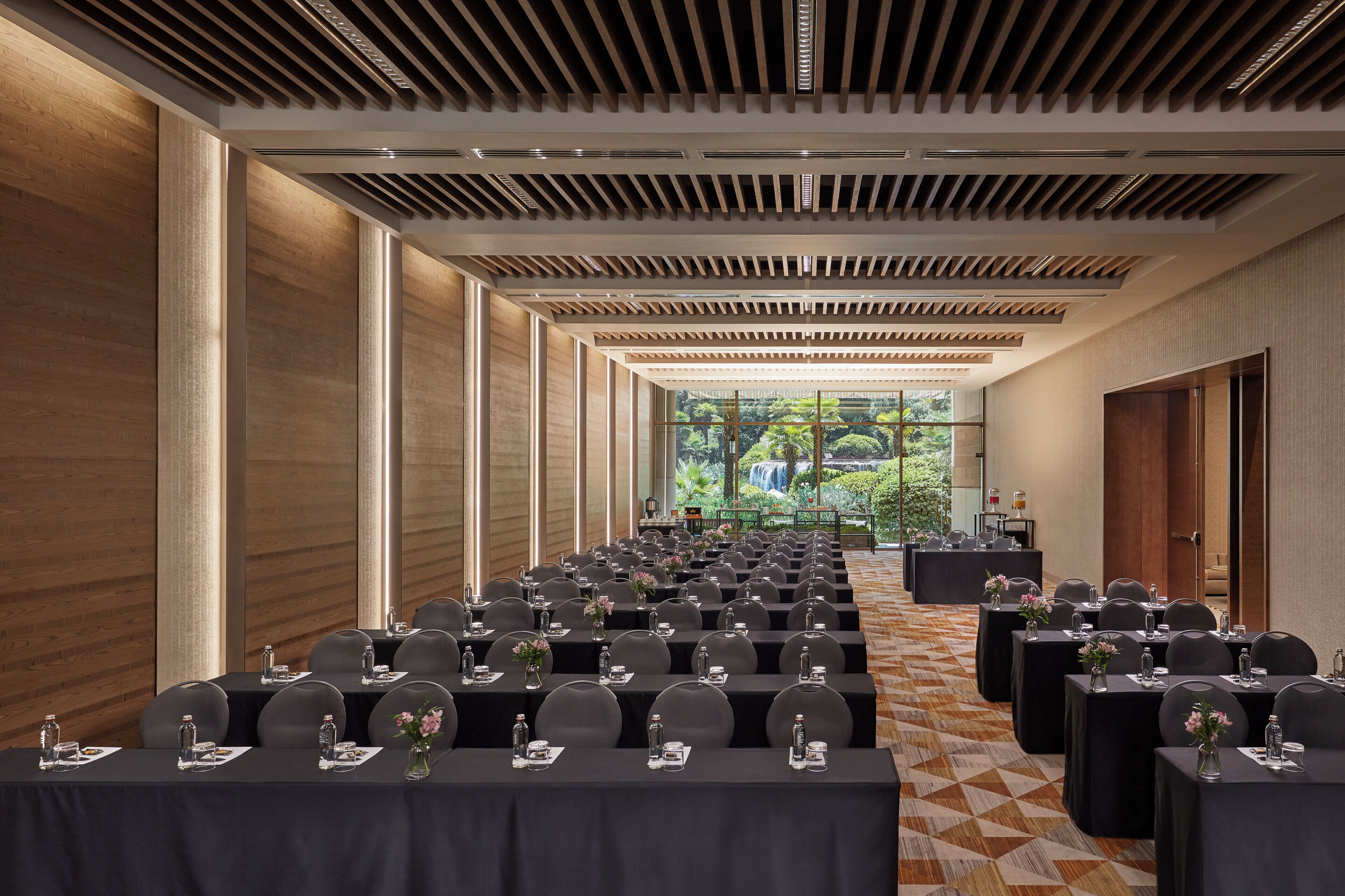A conference room with rows of tables and chairs, a large window with a view of greenery, and a modern ceiling design.