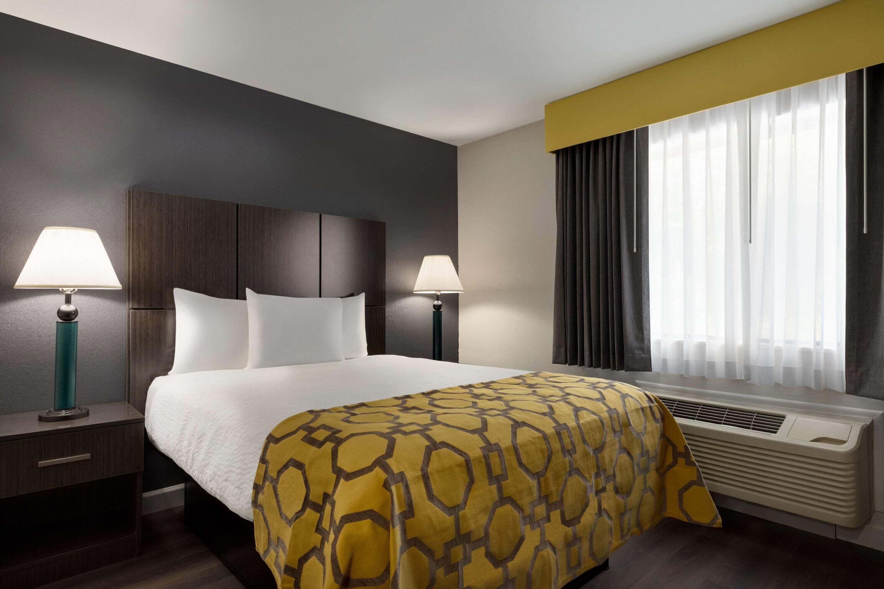 Camera d'albergo con un letto grande, due comodini con lampade, una finestra con tende e un condizionatore d'aria a parete.