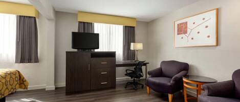 Habitación de hotel con cama, escritorio, televisor, dos sillones, una mesita y un cuadro en la pared.