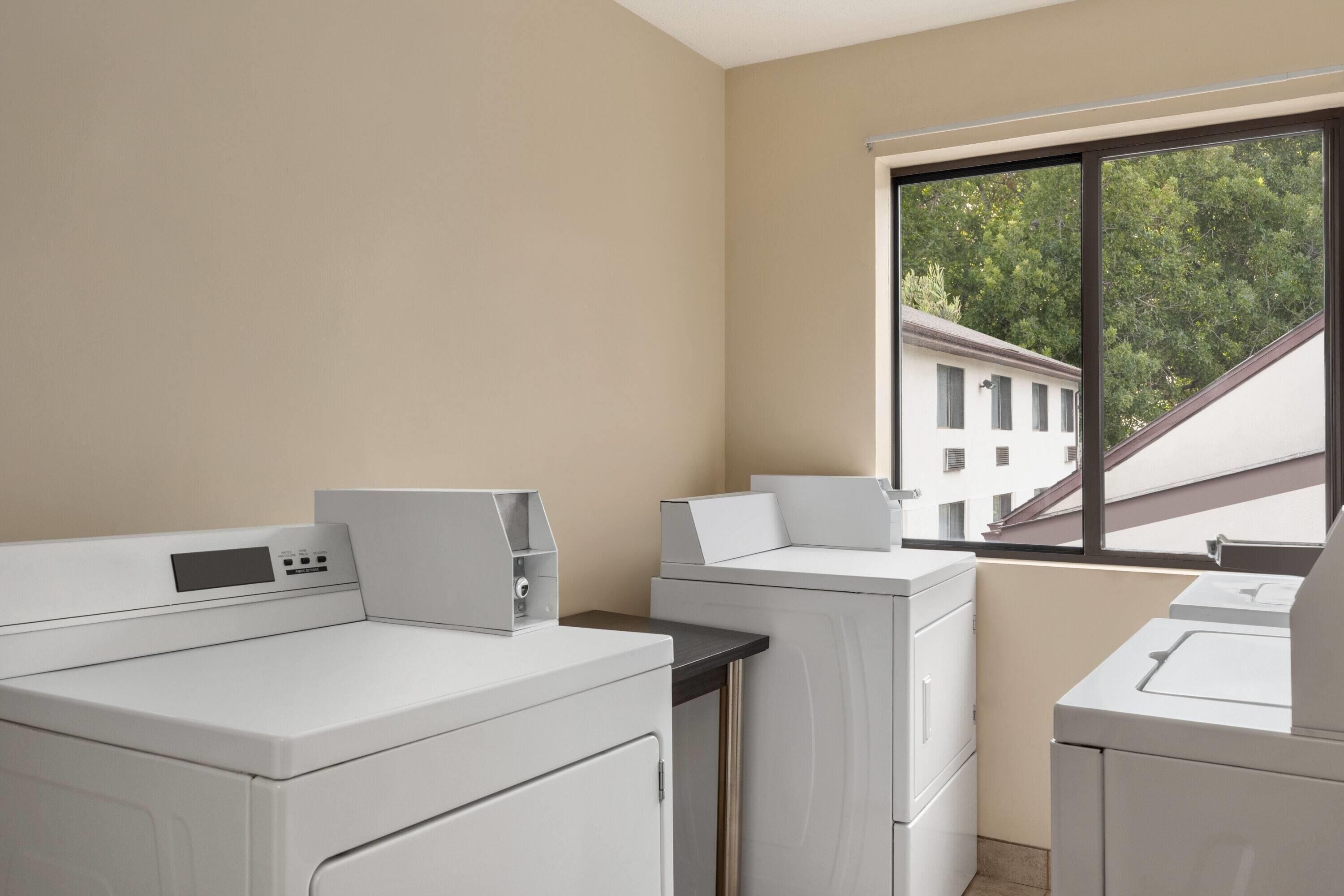 Een wasserette met gestapelde wasmachine en droger, een raam met uitzicht op gebouwen, en een beige muur.
