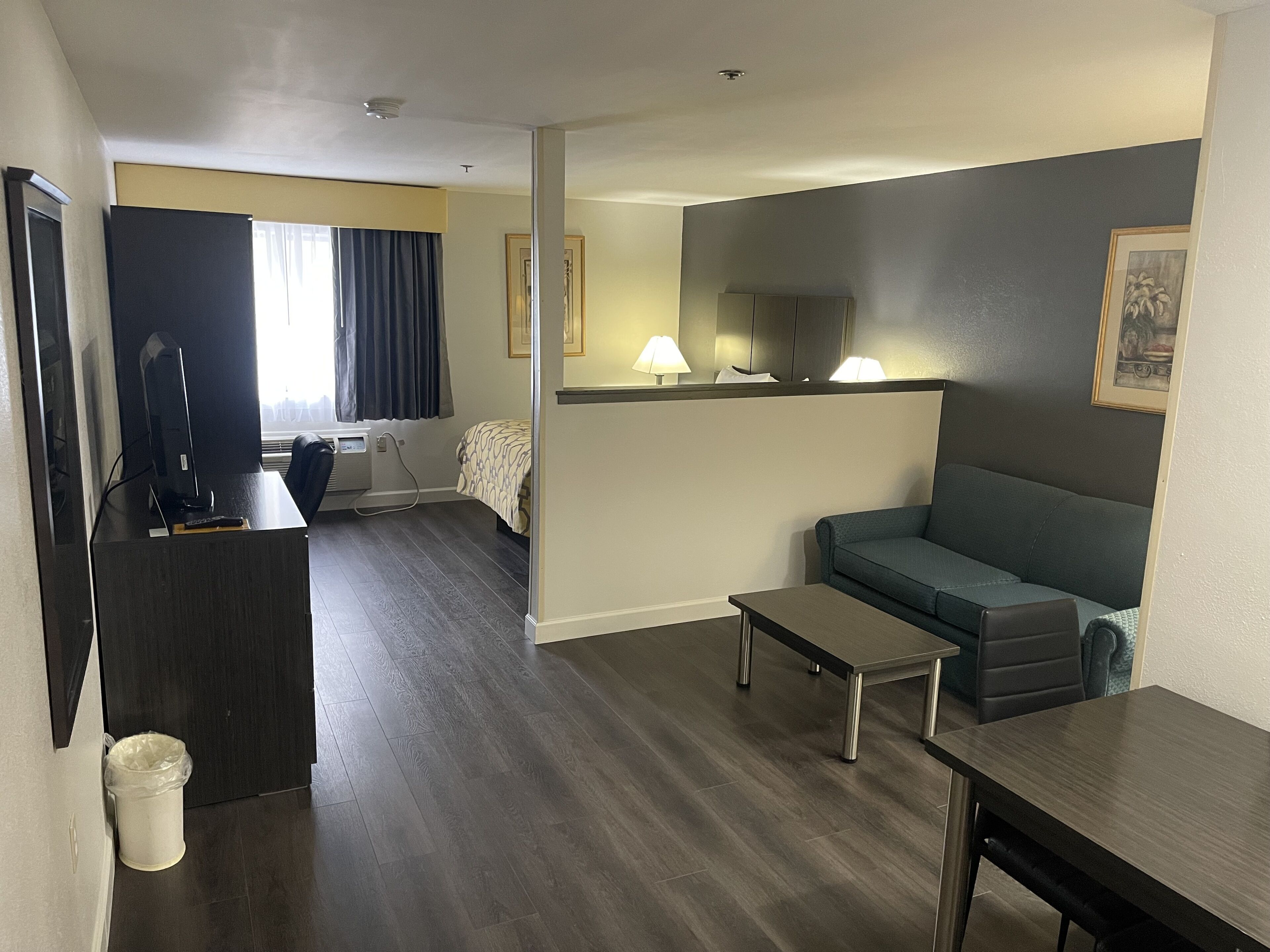 A modern hotel room with a dark wood floor, a grey sofa, a small table, a desk with a computer, and a bed with a patterned bedspread.