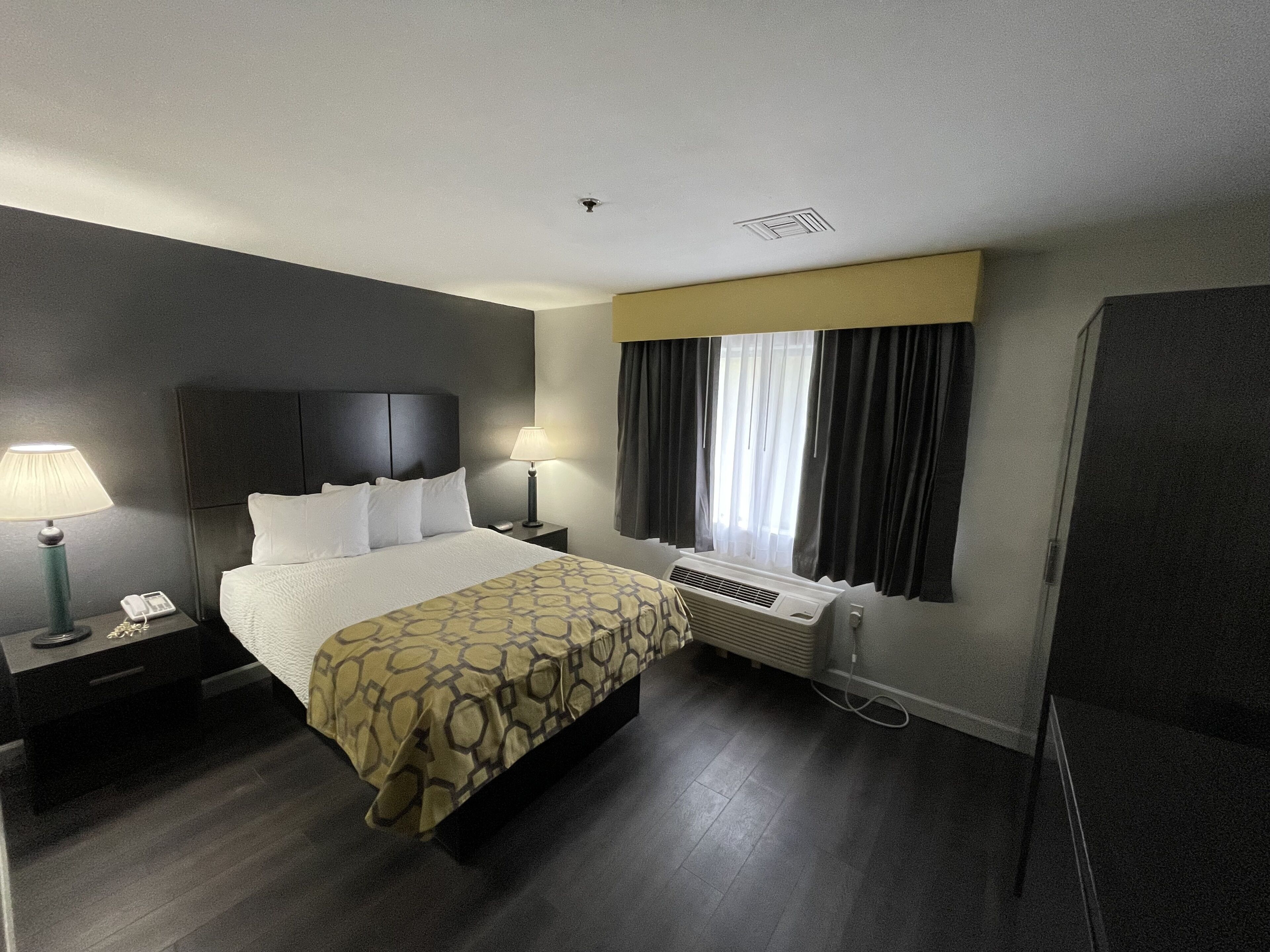 A hotel room with a bed, two bedside tables, a lamp, a telephone, a window with curtains, and a wall-mounted air conditioner.