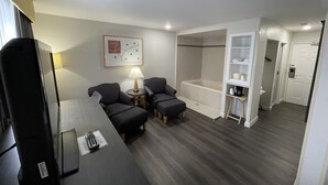 Ein Hotelzimmer mit einer Couch, zwei Sesseln, einem kleinen Tisch mit Fernbedienung, einer Lampe, einer Badewanne und einem begehbaren Kleiderschrank.