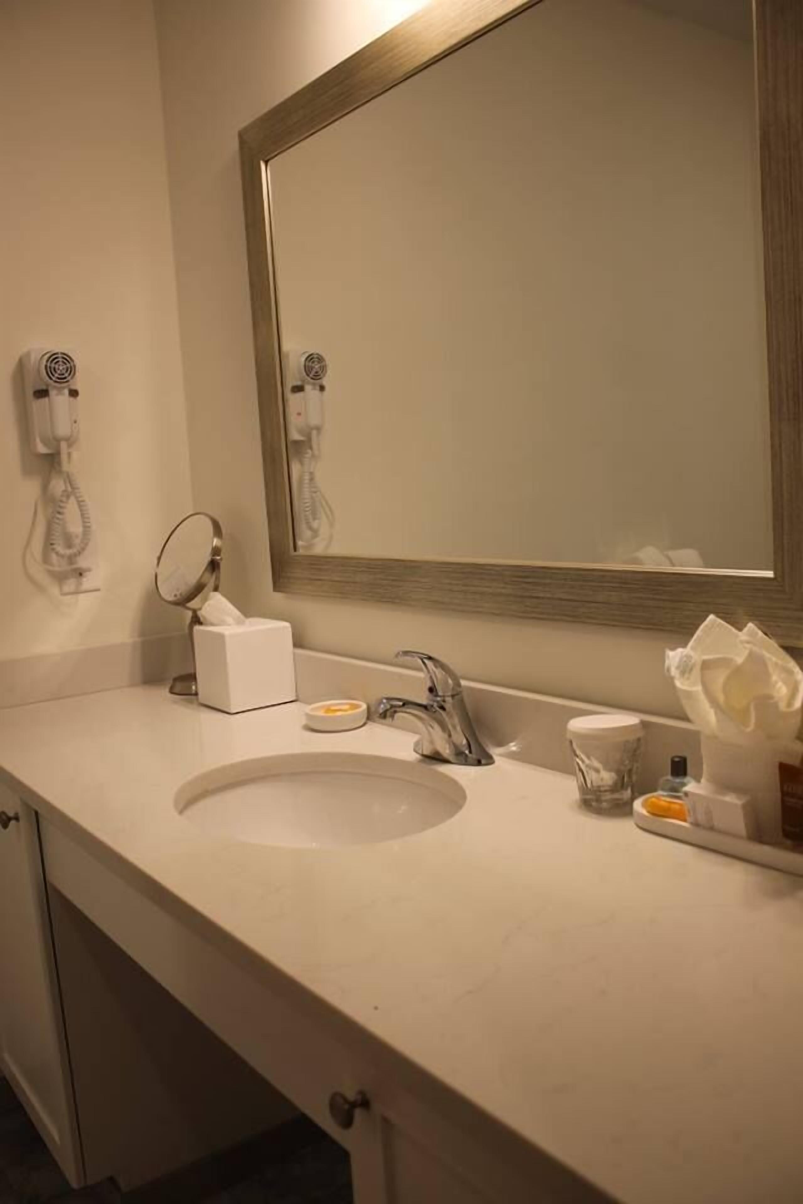 Casa de banho de hotel com um espelho grande, uma pia, um dispensador de toalhas e um secador de cabelo.