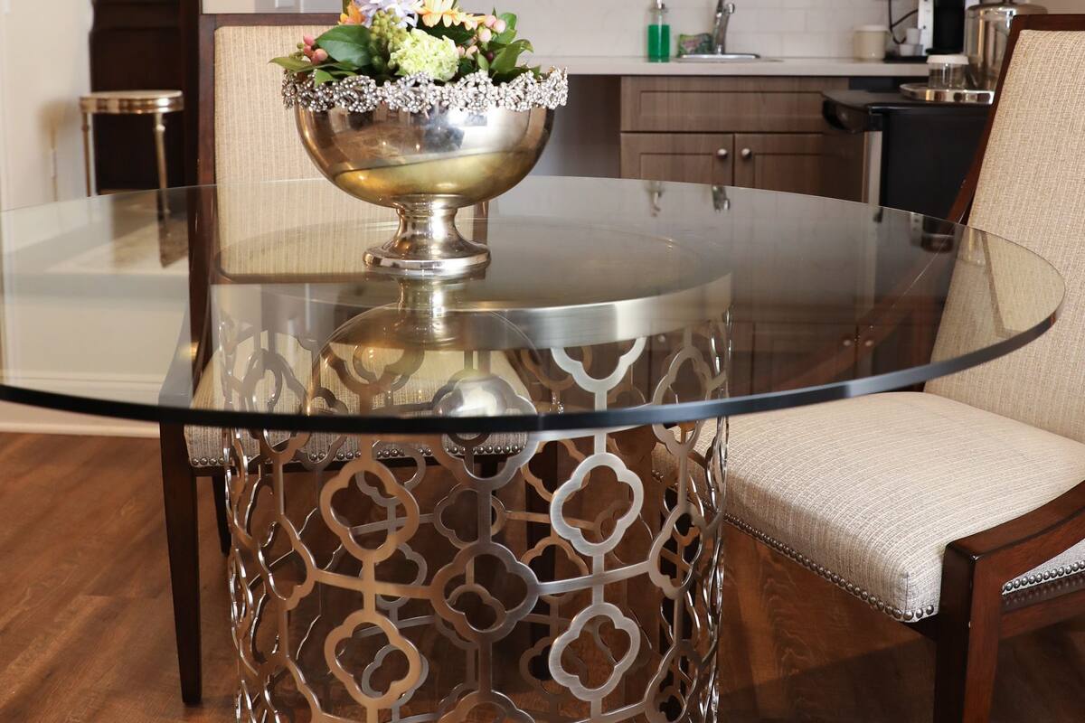 A glass-top dining table with a metal base, surrounded by a patterned metal chair.