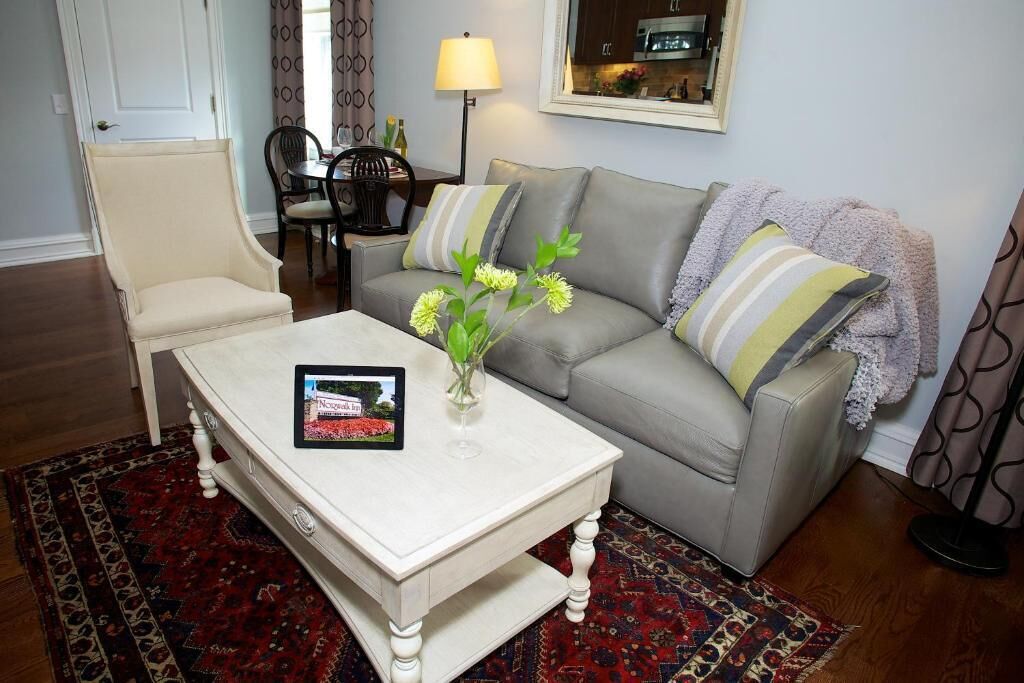 Ein Wohnzimmer mit einem beigefarbenen Sessel, einem grauen Sofa mit gestreiften Kissen, einem weißen Couchtisch mit Blumenvase und einem gemusterten Teppich.