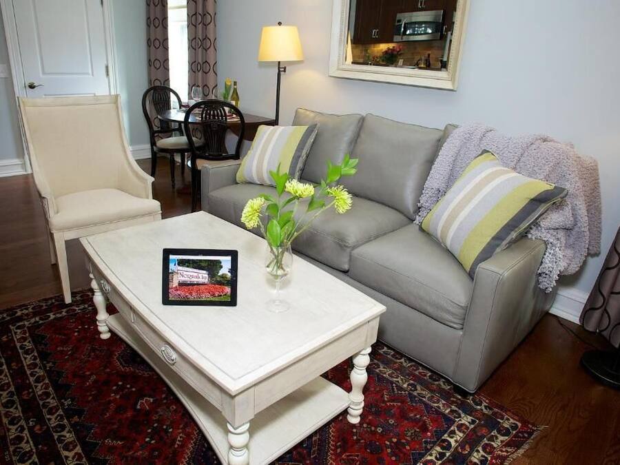 Ein Wohnzimmer mit einem beigefarbenen Sessel, einem grauen Sofa mit gestreiften Kissen, einem weißen Couchtisch mit Blumenvase und einem gemusterten Teppich.