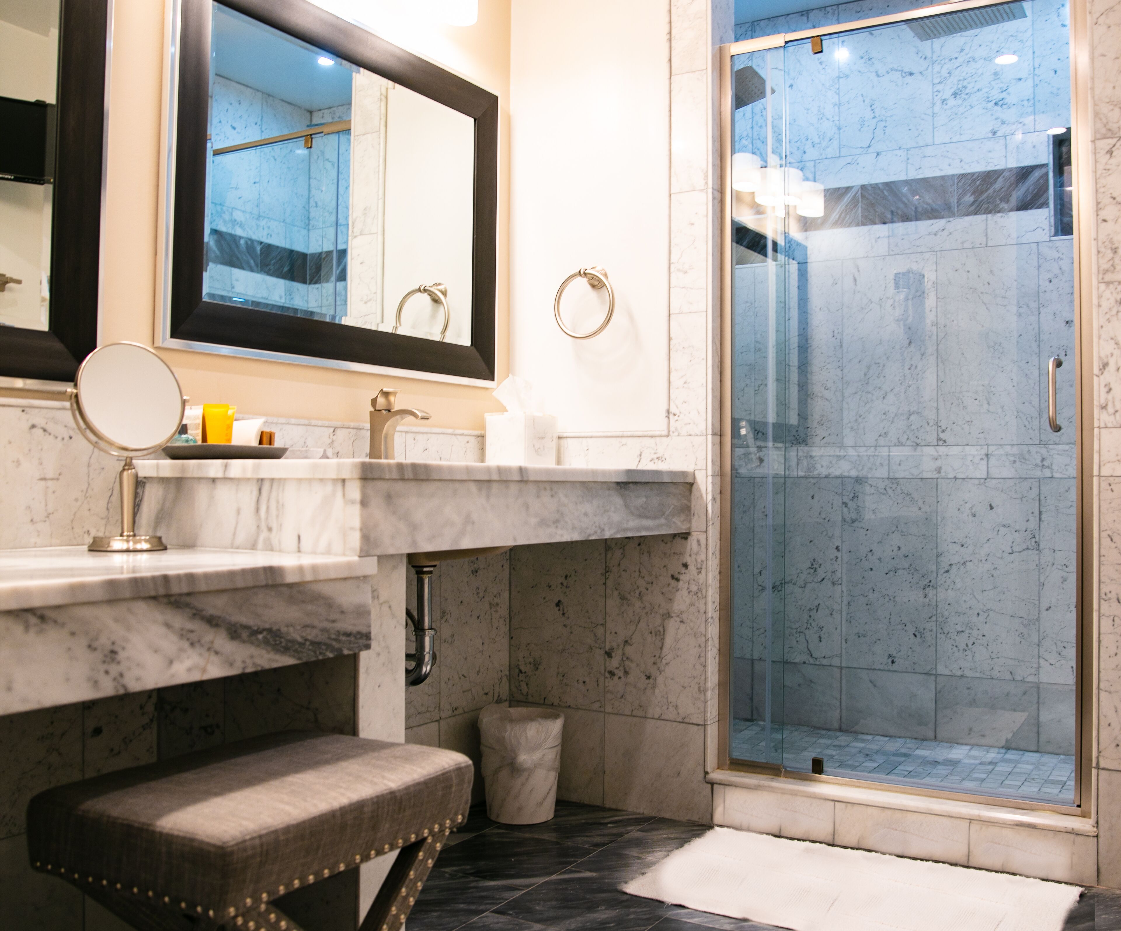 A bathroom with a marble sink, a round mirror, a bench, and a glass-enclosed shower.