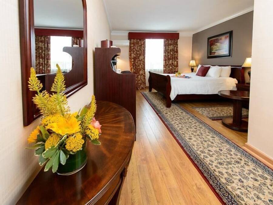 A hotel room with a bed, a desk with a mirror, a vase of flowers, and a patterned rug.