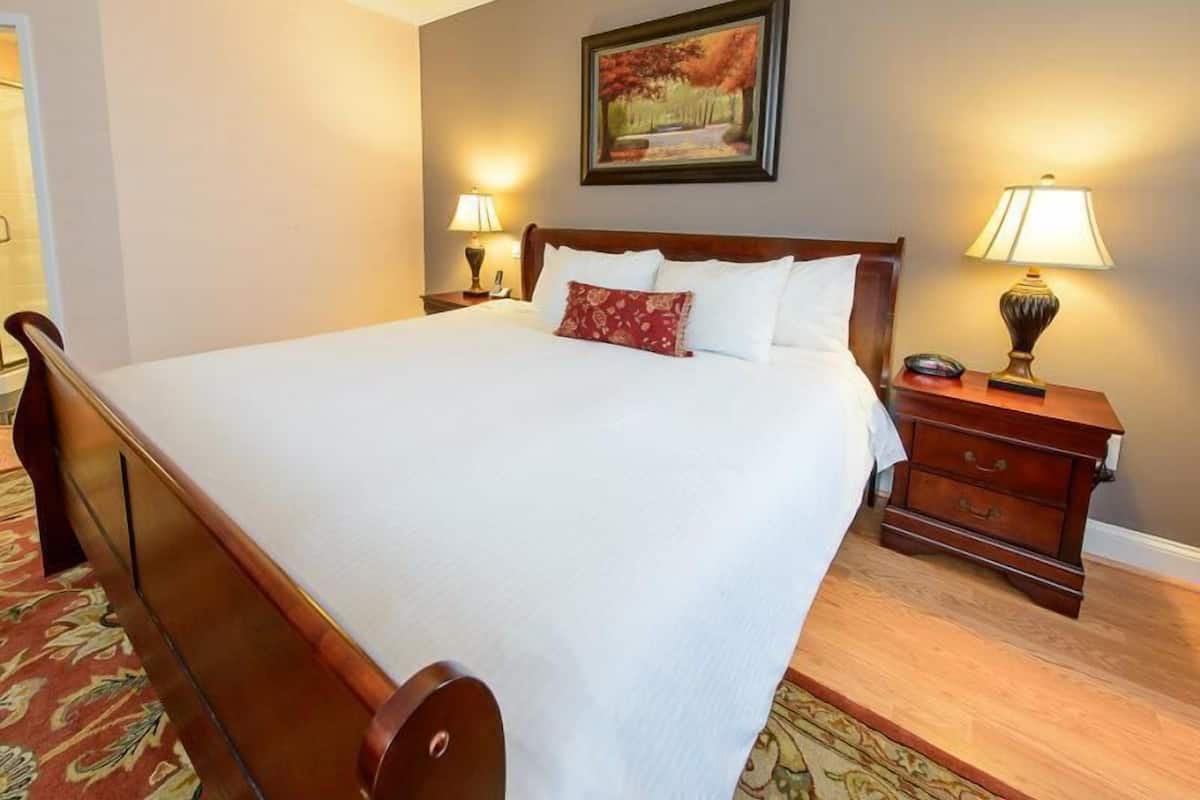 A neatly made bed with white linens and a red accent pillow, flanked by two bedside tables with lamps, in a room with a wooden floor and a patterned carpet.