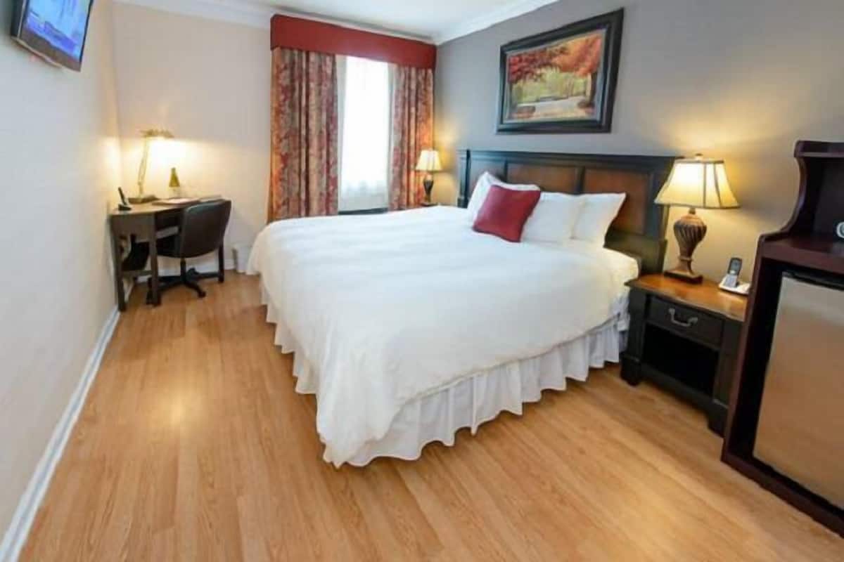 A hotel room with a large bed, a desk with a chair, a nightstand with a lamp, and a painting on the wall.