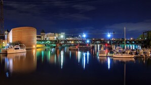 A waterfront marina at night with boats docked, a modern building with horizontal lines, and a bridge in the background.