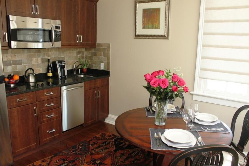 A kitchen with wooden cabinets, a stainless steel microwave, a sink, and a dining table set with plates, cutlery, and a vase of flowers.