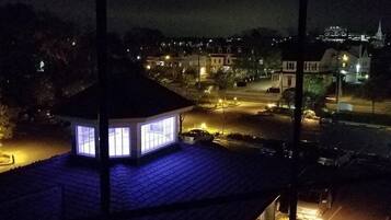 Vista nocturna de un edificio con una ventana iluminada, un área pavimentada y un horizonte urbano con edificios iluminados.