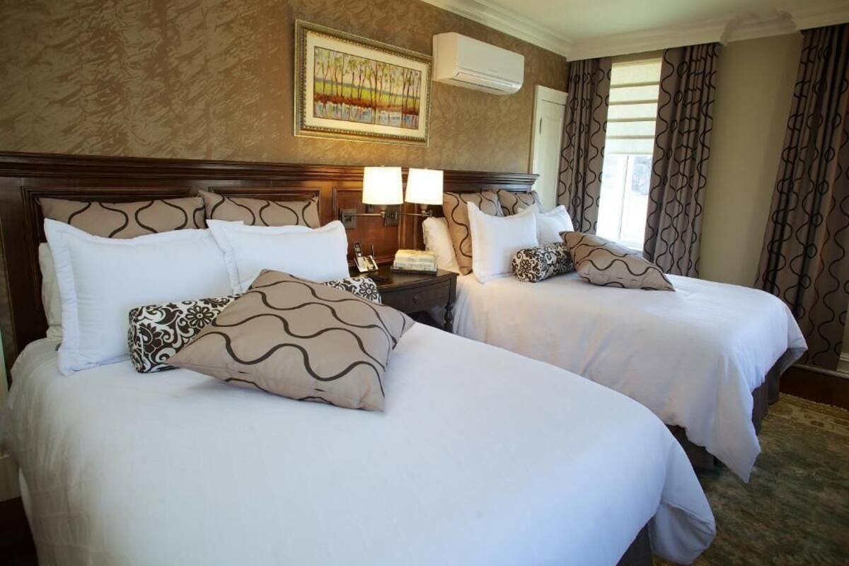 A hotel room with two beds, a nightstand with lamps, a painting on the wall, and a window with curtains.