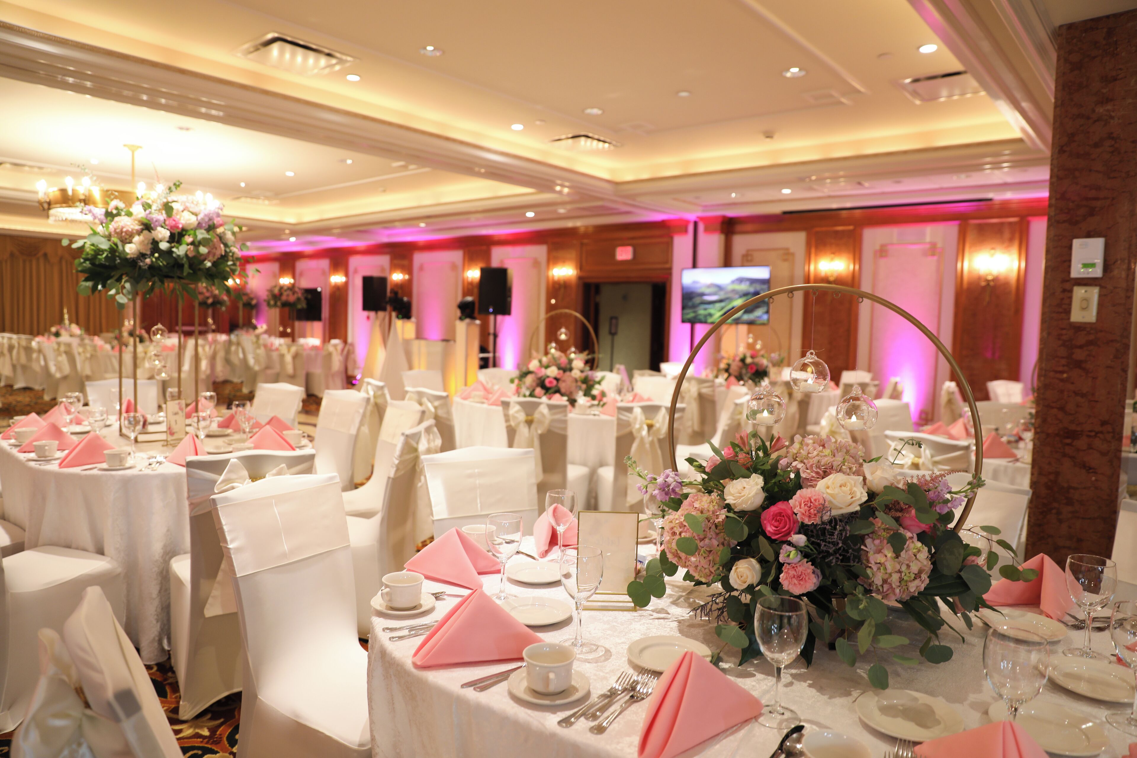Un salón de banquetes con mesas redondas dispuestas para un evento formal, con manteles blancos, servilletas rosas y centros de mesa florales.