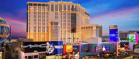 A large hotel with a prominent 'Planet Hollywood' sign, surrounded by various commercial buildings and illuminated billboards.