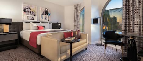 Habitación de hotel con cama, sofá, una mesita y una silla. Hay obras de arte en la pared y una ventana con cortinas.