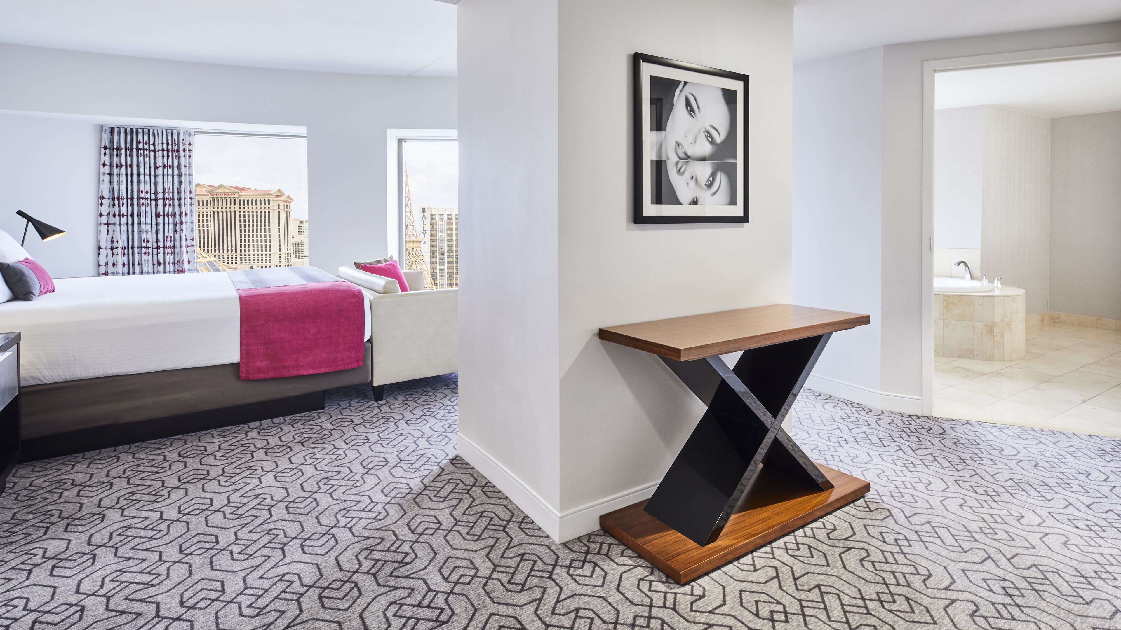 A modern hotel room with a bed, a bedside table, a window with a city view, and a framed picture on the wall.