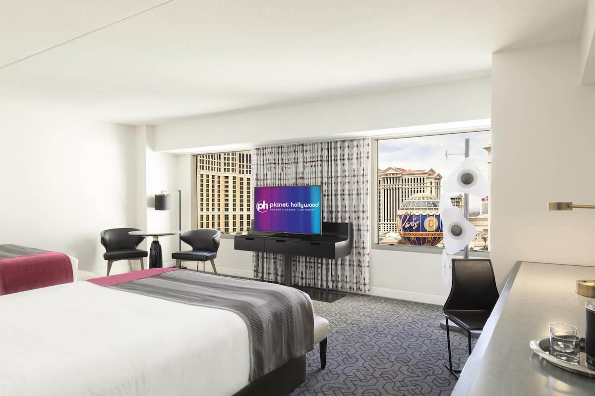 A hotel room with a bed, desk, chairs, a TV, and a view of a cityscape.