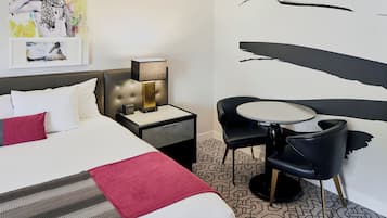 Una habitación de hotel con una cama, mesita de noche, lámpara y una pequeña mesa redonda con dos sillas.