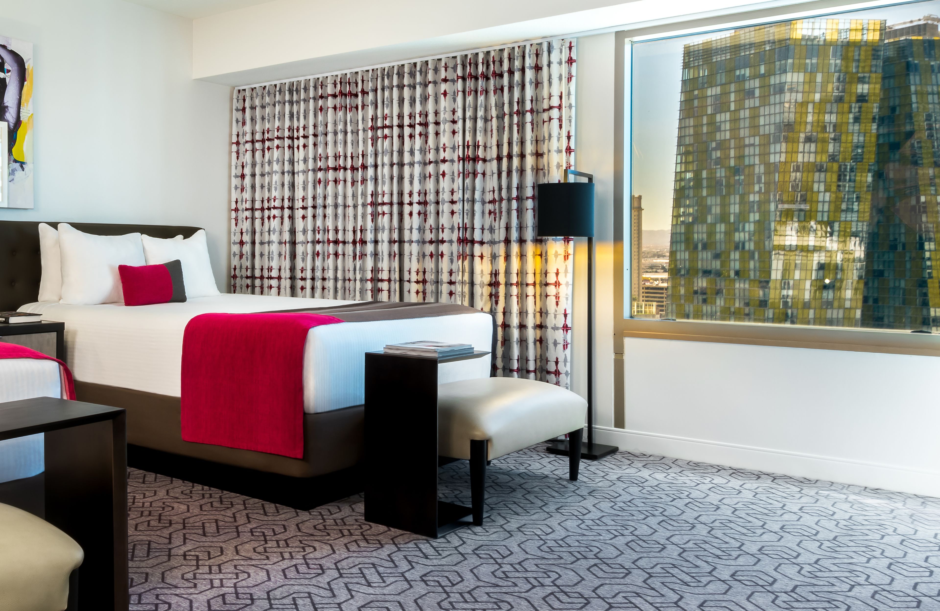 A hotel room with a bed, bedside tables, a bench, a lamp, and a large window with curtains.