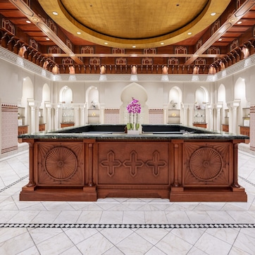 A spacious lobby with a central marble reception desk, high wooden ceilings, and arched white columns.