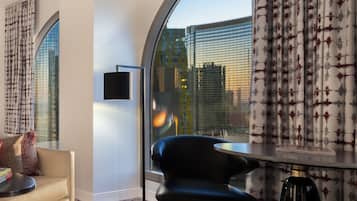 A modern hotel room with a curved window offering a city view, a black chair, a small table, and a floor lamp.