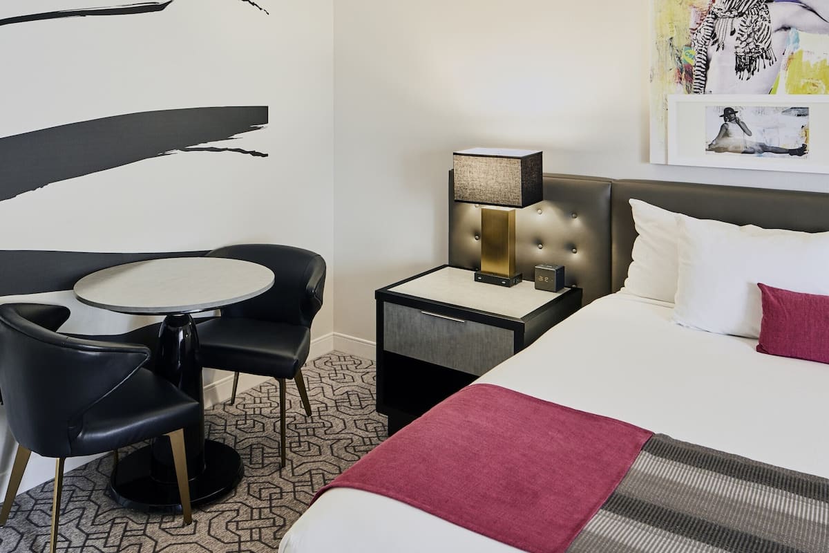 A hotel room with a bed, a bedside table with a lamp, a small round table with two chairs, and a large abstract painting on the wall.