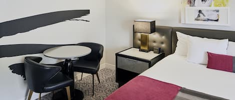 A hotel room with a bed, a bedside table with a lamp, a small round table with two chairs, and a large abstract painting on the wall.