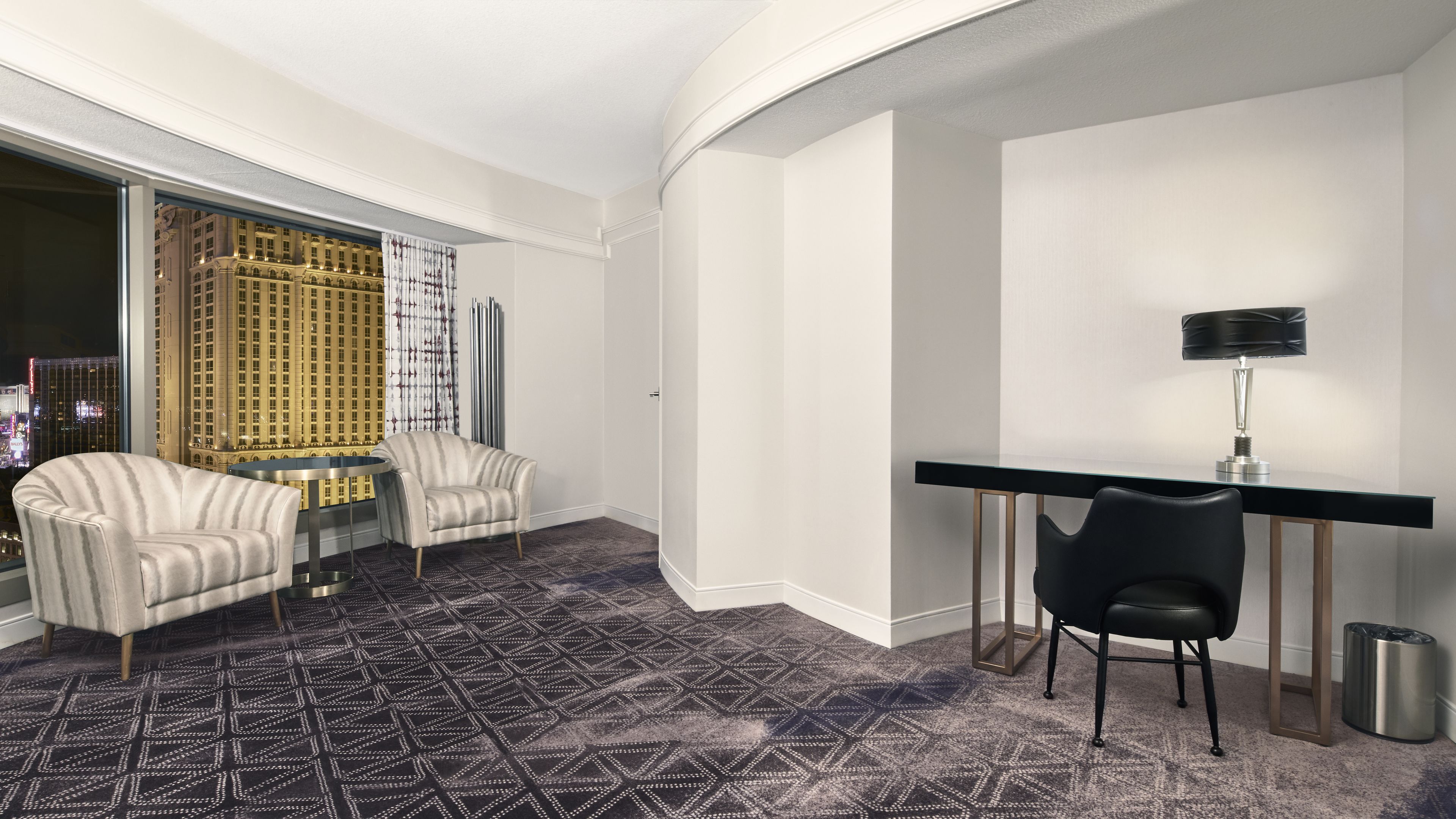 Habitación de hotel con un ventanal grande que ofrece vista a la ciudad, una mesa redonda, dos sillones, un escritorio con una silla y una alfombra estampada.