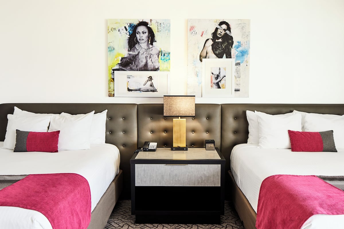 A hotel room with two beds, a nightstand with a lamp, and artwork on the wall.