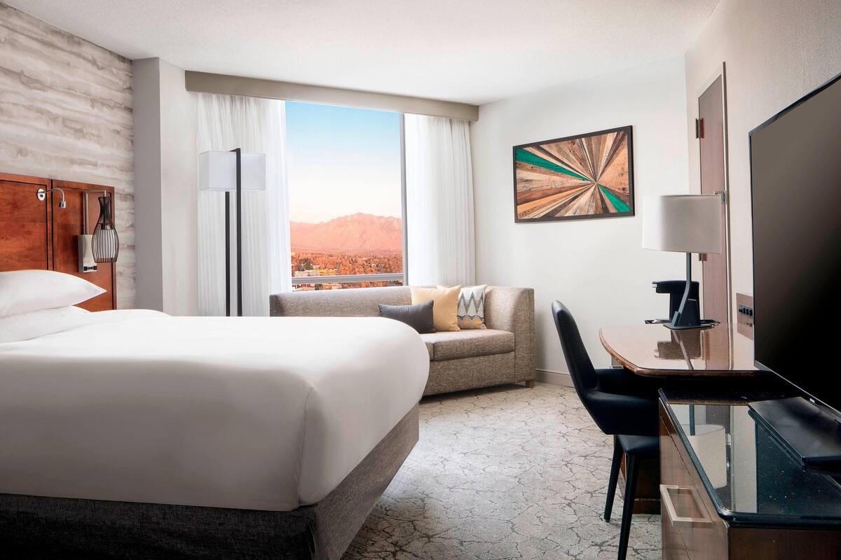 A hotel room with a large bed, a desk, a chair, a TV, and a view of mountains.