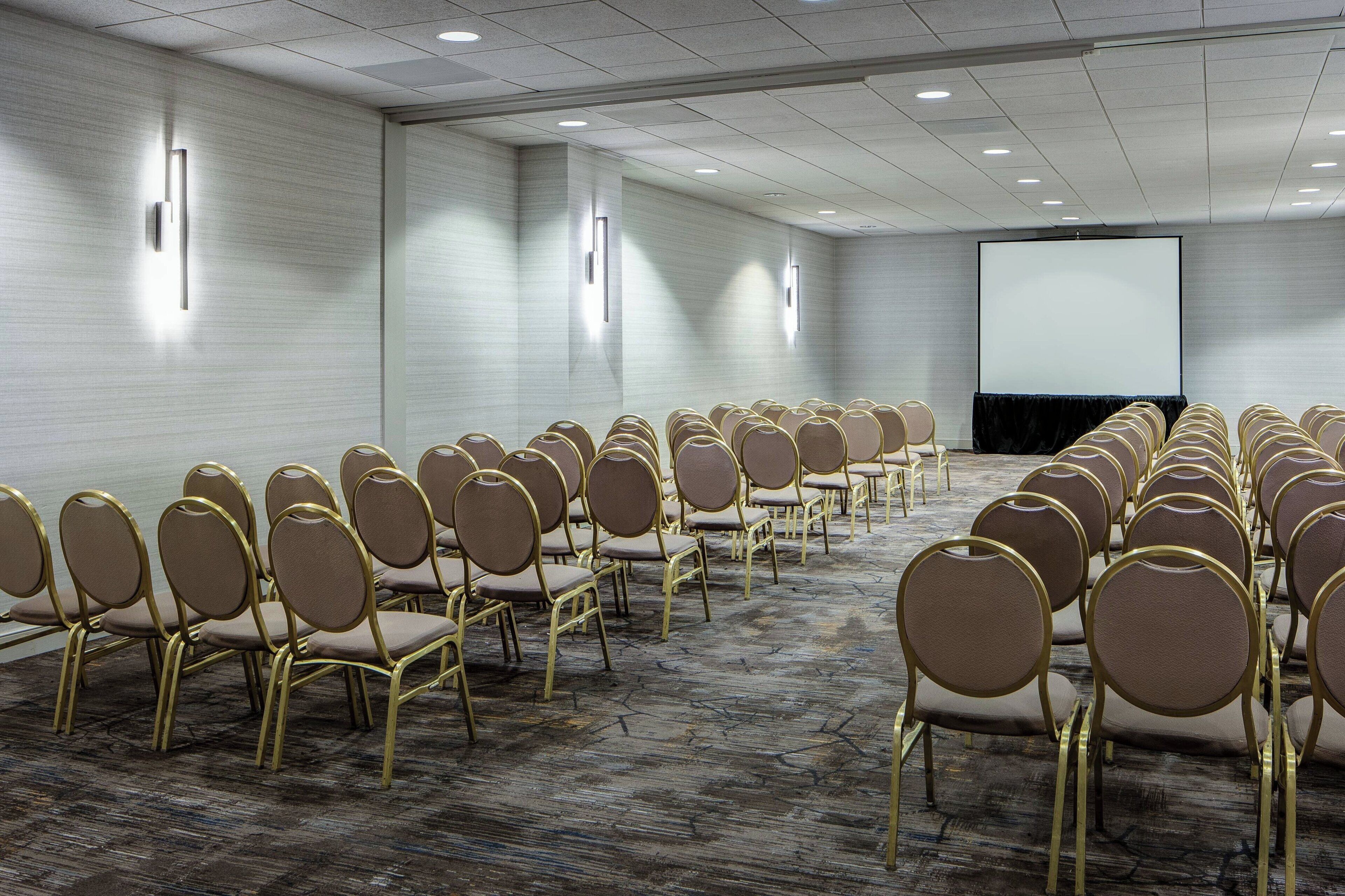 A conference room with rows of chairs, a projector screen, and wall-mounted lighting.