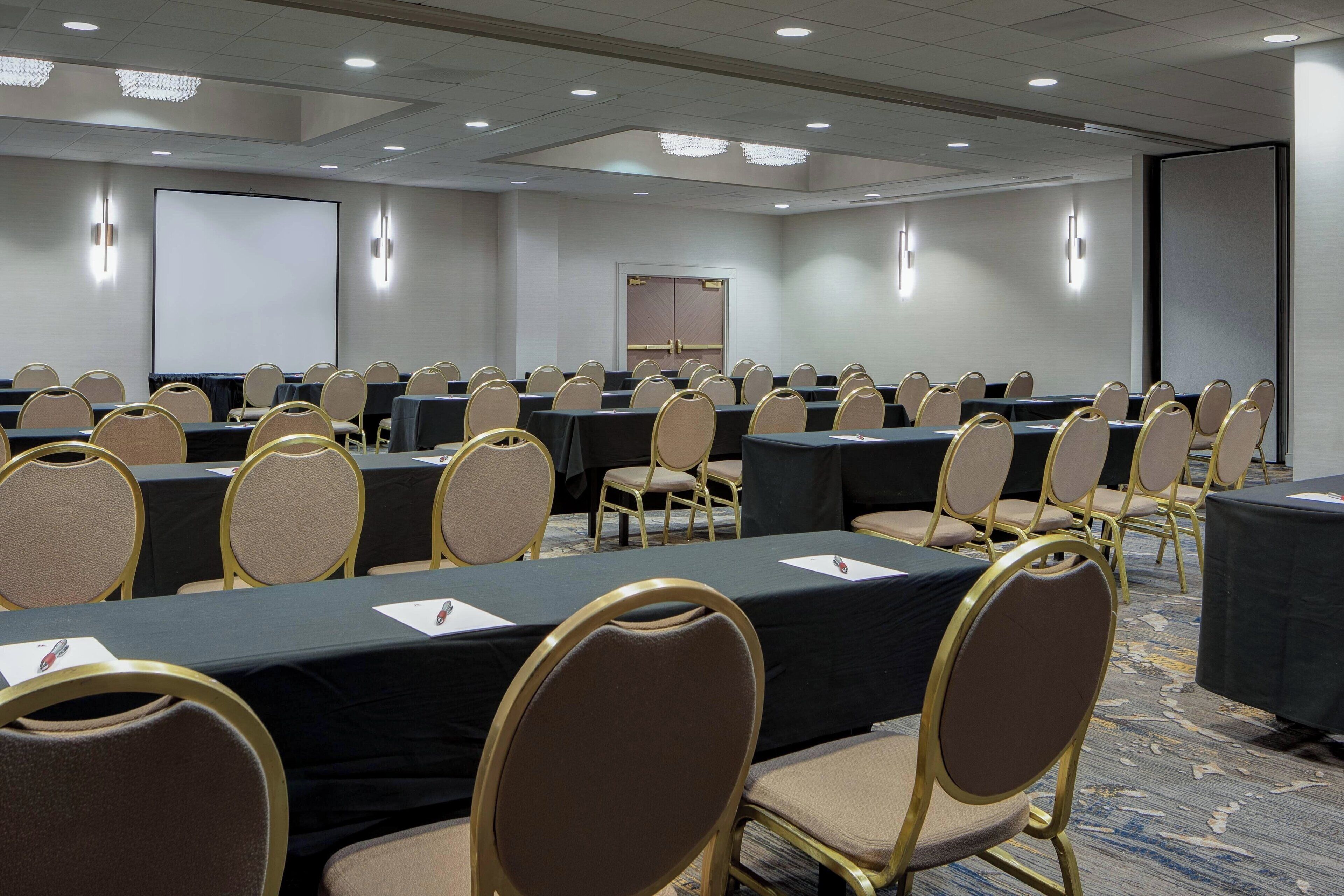 A conference room with rows of tables and chairs, a projector screen, and a door in the background.