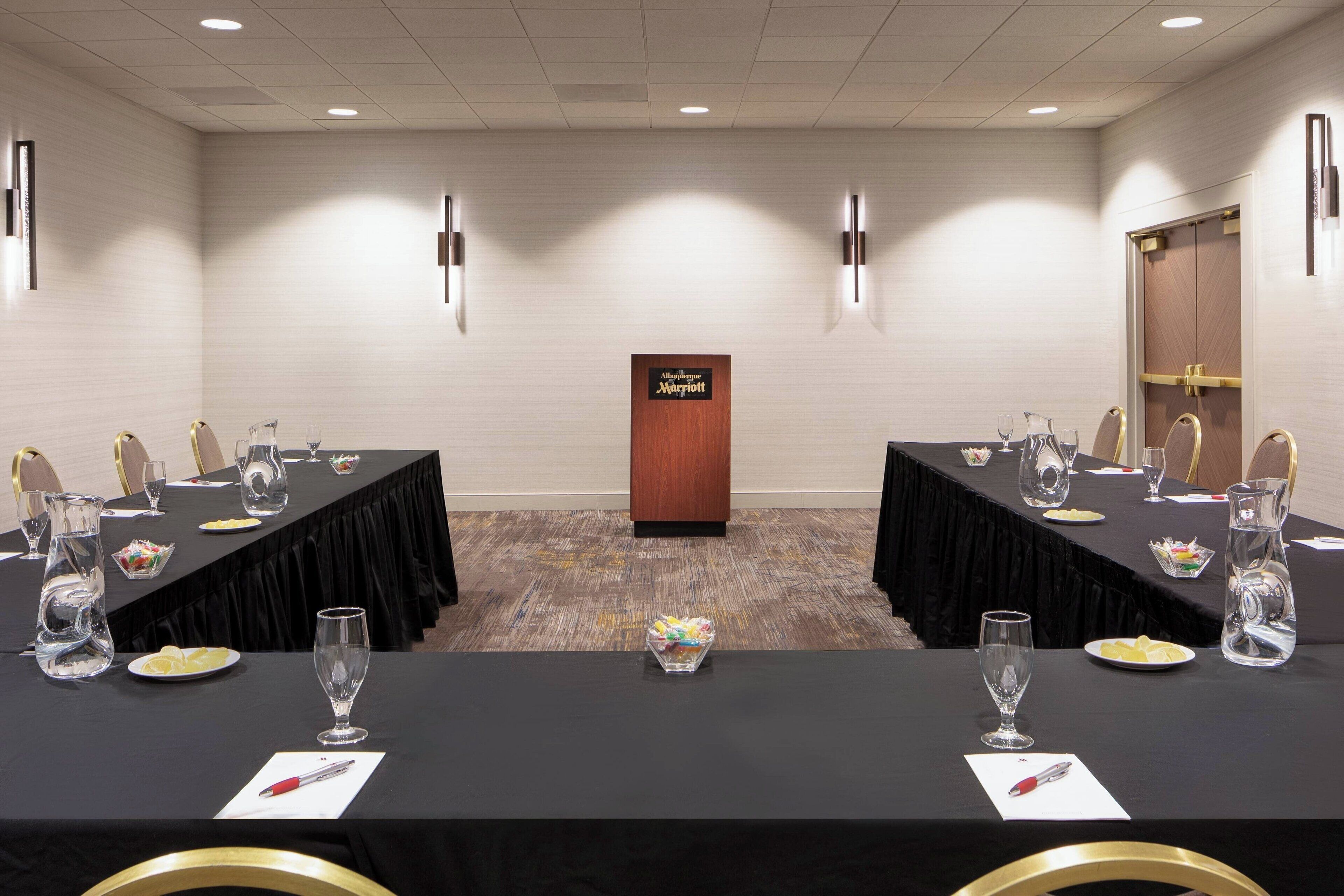 A conference room with two long tables, each with chairs, glassware, and a pitcher. A wooden podium with the Marriott logo is in the center.