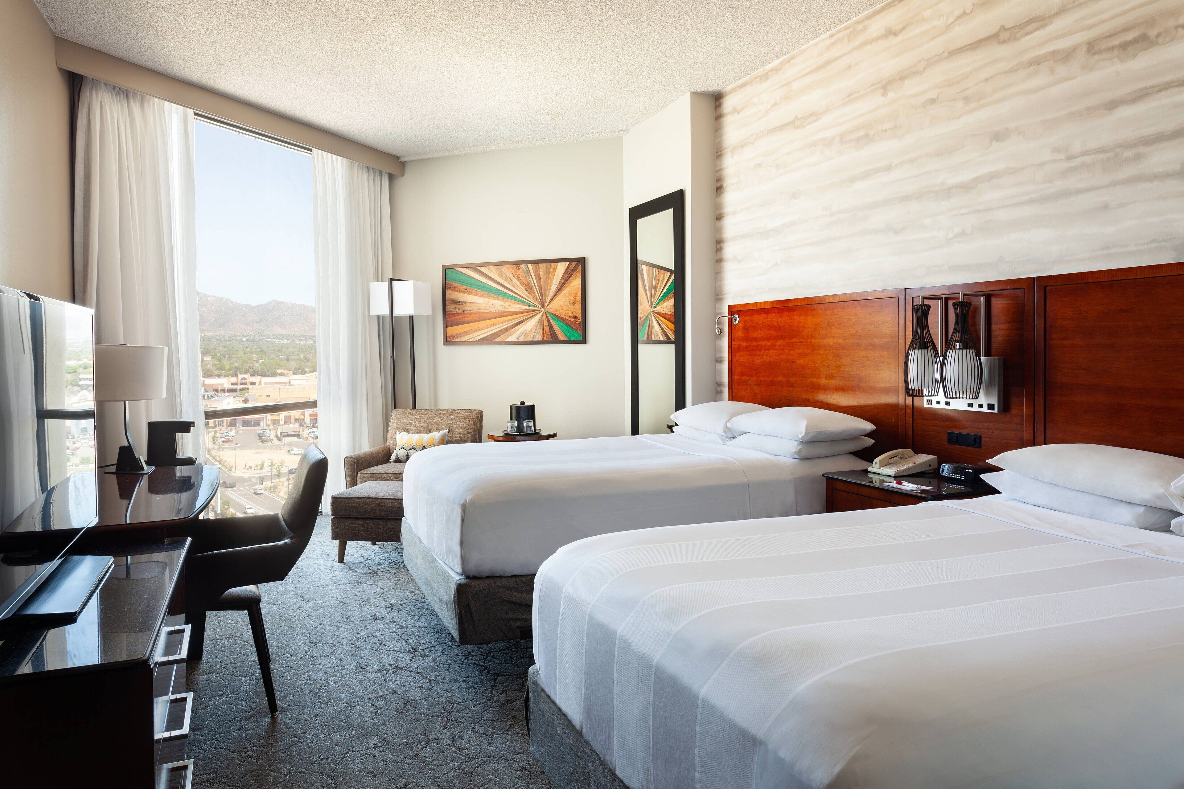 A hotel room with two beds, a desk, a chair, a TV, and a view of the city.
