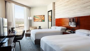 A hotel room with two beds, a desk, a chair, a TV, and a view of the city.