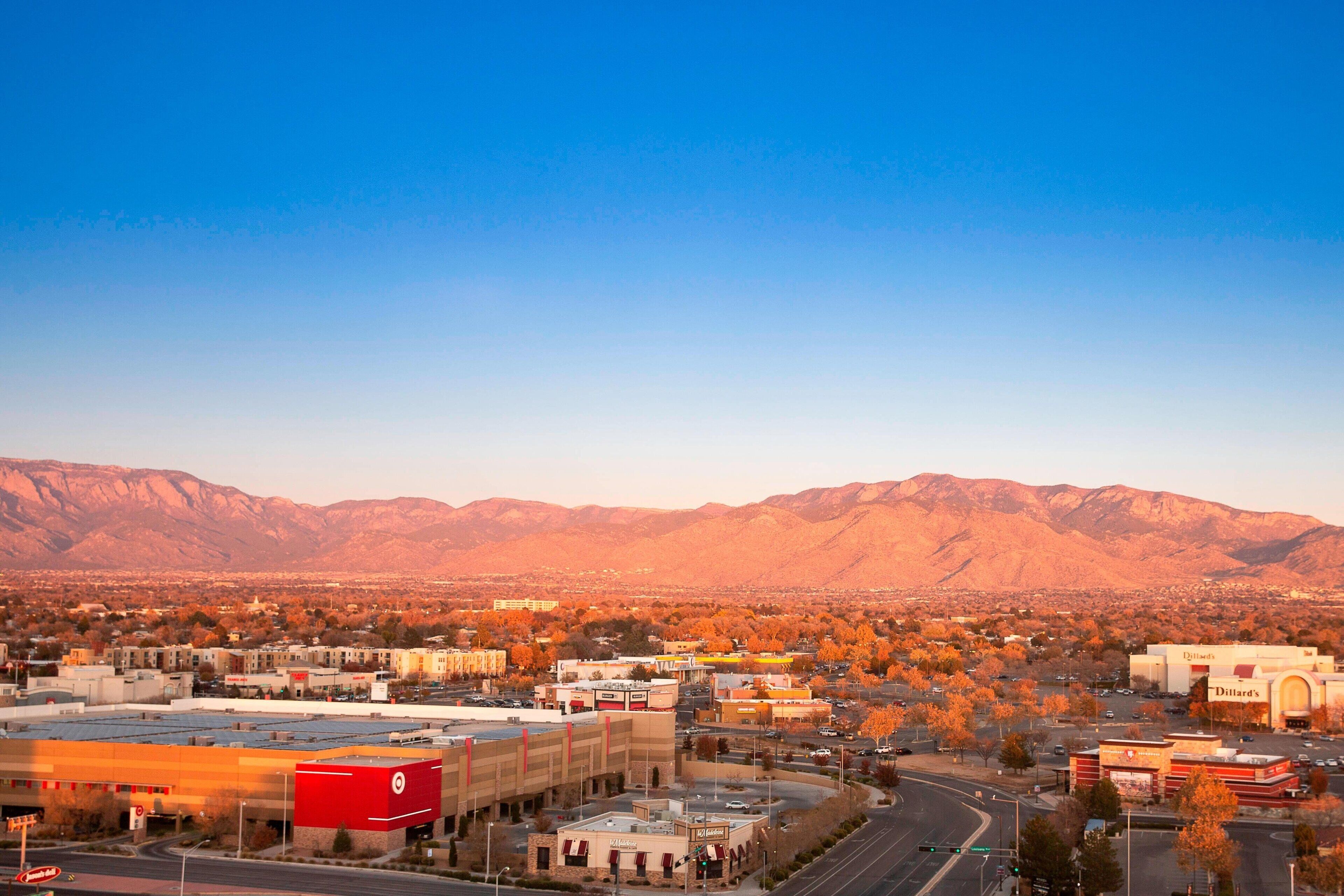A cityscape with a prominent building featuring a red and white sign, surrounded by autumn-colored trees and mountains in the background.