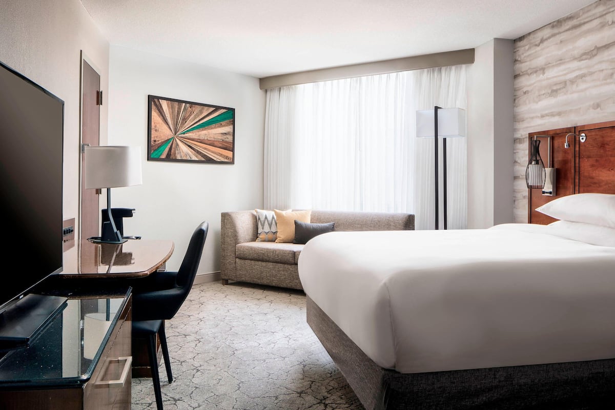 A hotel room with a large bed, a desk with a chair, a sofa, a TV, and a painting on the wall.