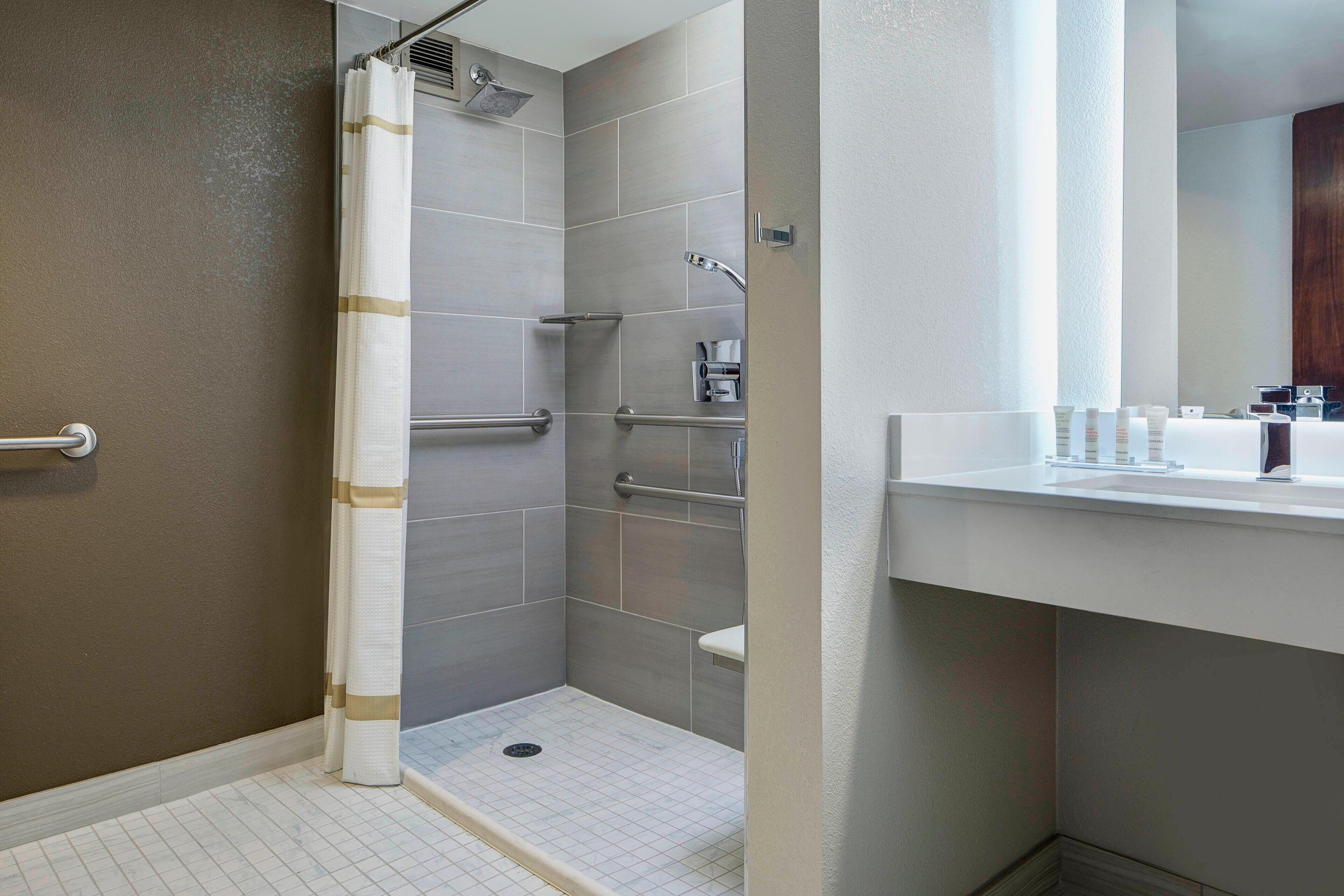 Un baño moderno con ducha, barras de apoyo y un lavamanos blanco.