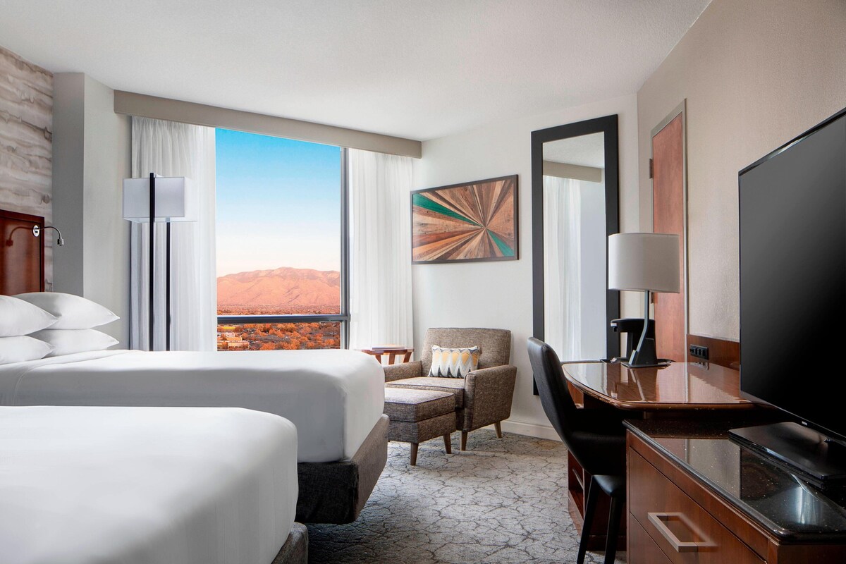 Habitación de hotel con dos camas, un escritorio, una silla, un televisor y un ventanal grande que ofrece vistas al desierto.