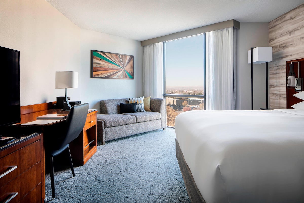 A hotel room with a large bed, a desk with a computer, a sofa, and a large window with a city view.